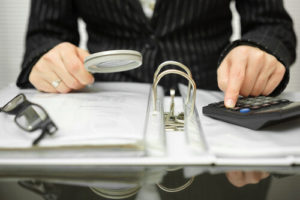 Person in suit using calculator while examining documents in binder with magnifying glass
