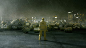Worker in HazMat suit standing in front of pile of barrels in rain