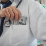 doctor in Frame from neck down to chest in white coat putting hundred dollar bills in his coat pocket to represent taking money for himself