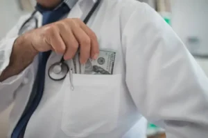 doctor in Frame from neck down to chest in white coat putting hundred dollar bills in his coat pocket to represent taking money for himself