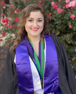 Brunette woman smiling in black graduation gown with purple sash standing in front of a red rose bush.